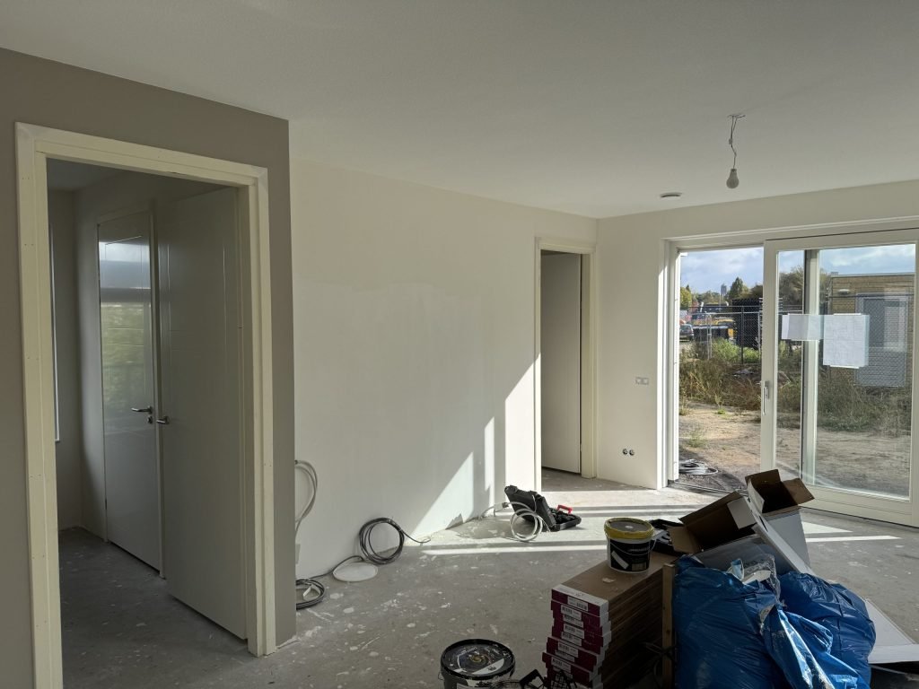 Nieuwbouwwoonkamer tijdens de afwerking, met dozen laminaat, verfemmers en een schuifpui