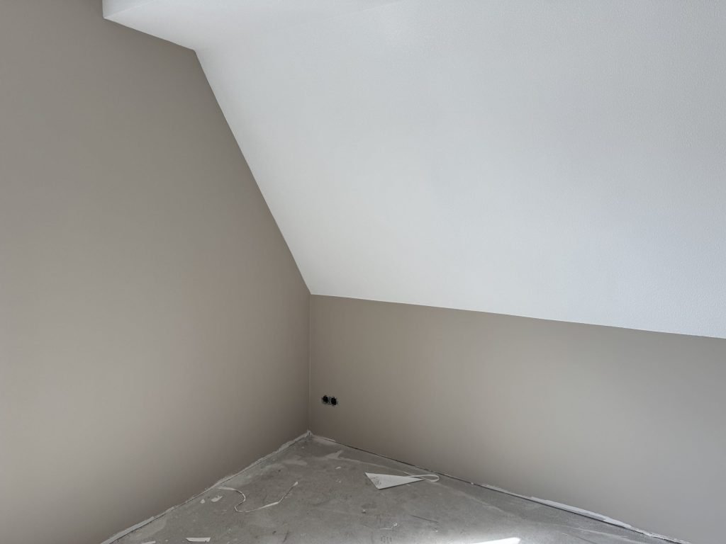 Zolderkamer met een schuine witte wand en beige gesausde muren op een betonvloer
